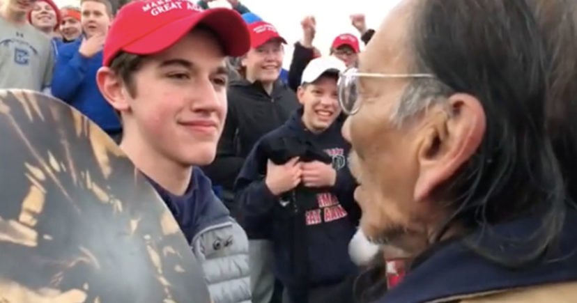 A teenager wearing a Make America Great Again hat stands in front of an elderly Native American singing and playing a drum in Washington